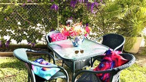 A small outdoor seating area with a table and two chairs, surrounded by lush greenery and purple flowers. - Villa Mia Apartment Studio #4 (Oistins)