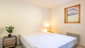 A neatly made bed in a room with a small bedside table, a lamp, and a view of a landscape through a window. - Apartment in Saint-Sorlin near Ski Resort (Saint-Sorlin-d´Arves)