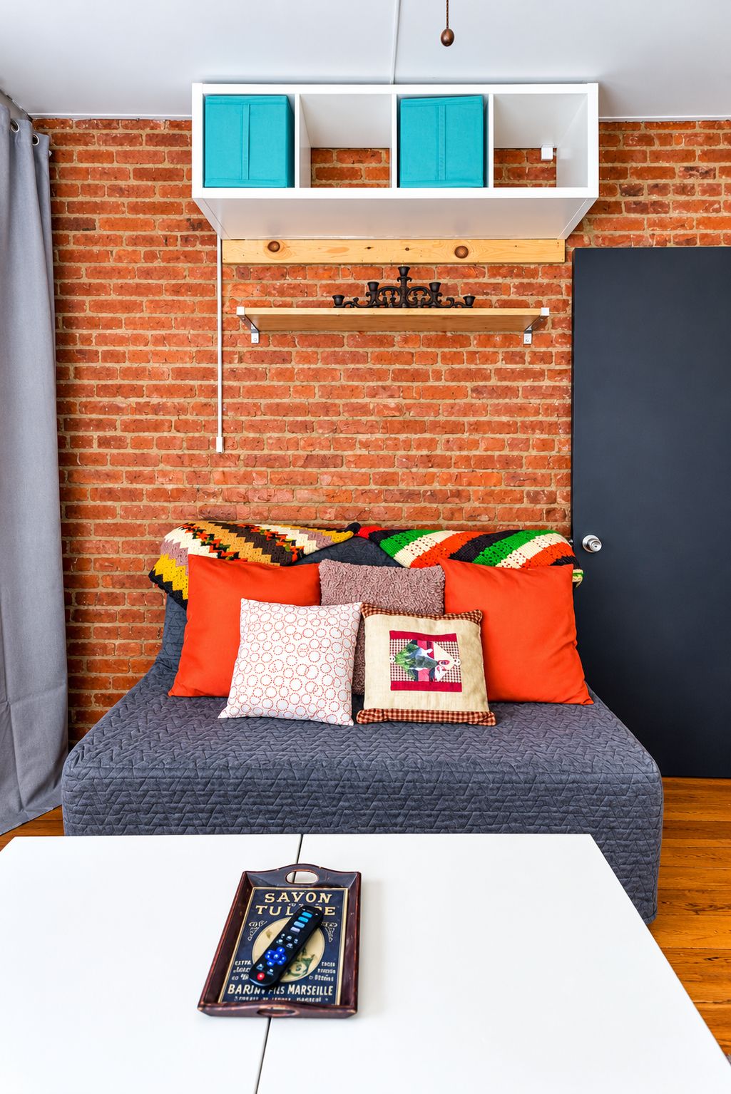 A room with a brick wall, a bed with multiple pillows, a white table with a remote control, and a door.