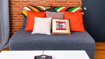 A room with a brick wall, a bed with multiple pillows, a white table with a remote control, and a door.