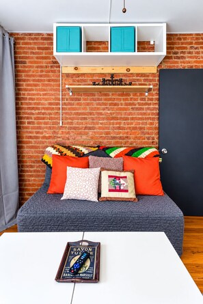 A room with a brick wall, a bed with multiple pillows, a white table with a remote control, and a door.
