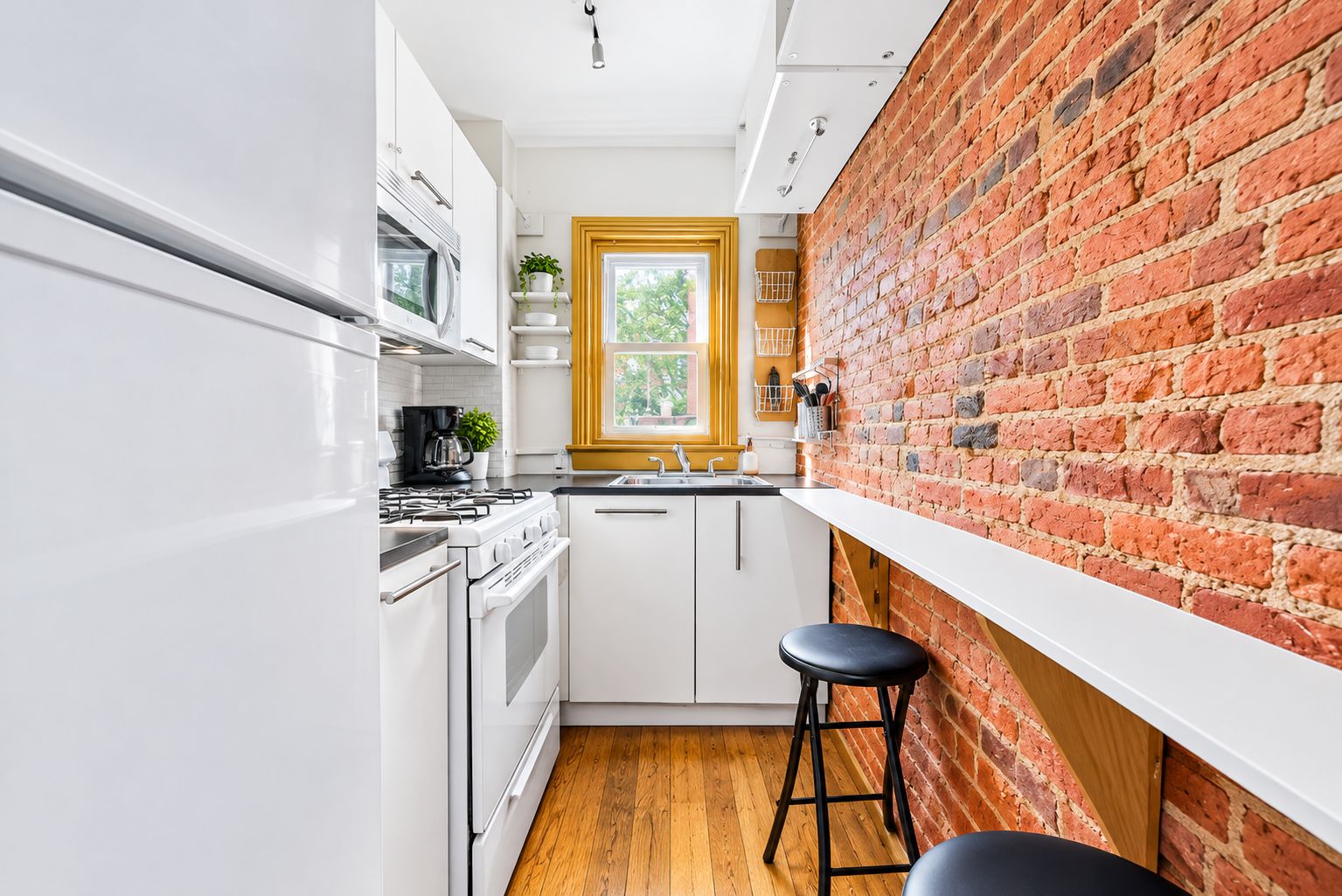 A compact kitchen with white cabinetry, a brick accent wall, a window with a yellow frame, and a wooden floor.