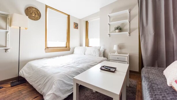 Una habitación de hotel moderna con una cama grande, un escritorio blanco, una mesita de noche, un estante con una planta y una ventana con cortinas.