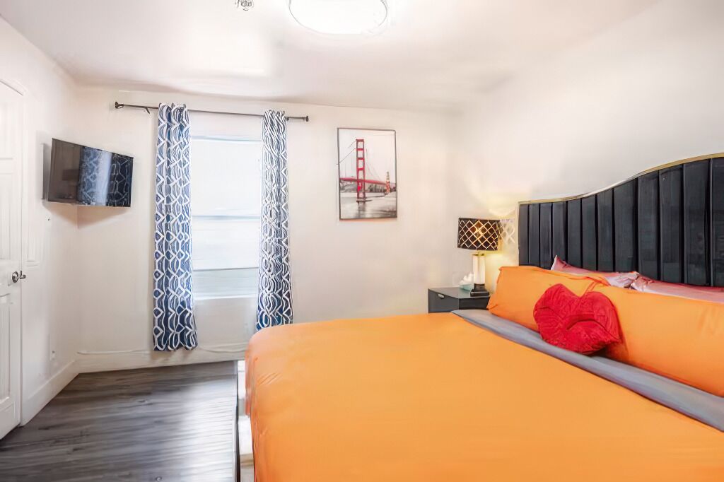 A bedroom with a bed, a TV mounted on the wall, a nightstand, and a window with curtains.