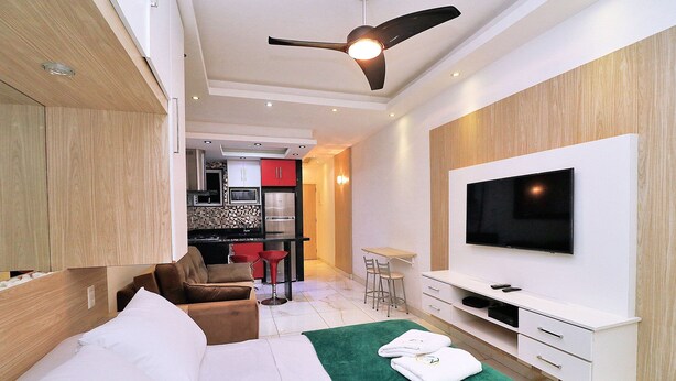 A modern hotel room with a bed, a green area rug, a TV, a ceiling fan, and a kitchenette in the background. - Prado X (Rio de Janeiro)