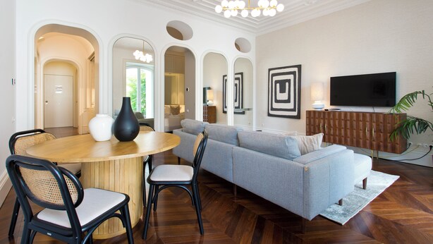 Un salon moderne avec une table ronde en bois, deux chaises noires, un canapé gris et un téléviseur posé sur un meuble en bois.