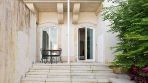 Un escalier en pierre mène à une porte dotée d’un panneau de verre, une petite table avec des chaises se trouve à proximité, et un arbuste vert est placé sur la droite.
