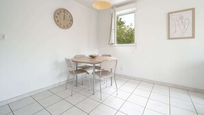 A dining area with a round table, four chairs, a clock, a window with a view, and a framed artwork on the wall. - Coastal Comfort for Four (Le Grau-du-Roi)