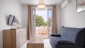 A modern apartment with a grey sofa, wooden cabinets, a small table, and a TV on a cabinet. - Coastal Comfort for Four (Le Grau-du-Roi)