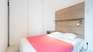 A bedroom with a bed, a wooden headboard, a nightstand, and a wall-mounted light fixture. - Coastal Comfort for Four (Le Grau-du-Roi)