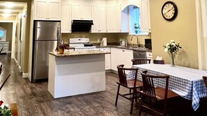 Une cuisine moderne avec des meubles blancs, un réfrigérateur en acier inoxydable, un four encastré et un coin repas agrémenté d’une nappe à carreaux et de chaises.