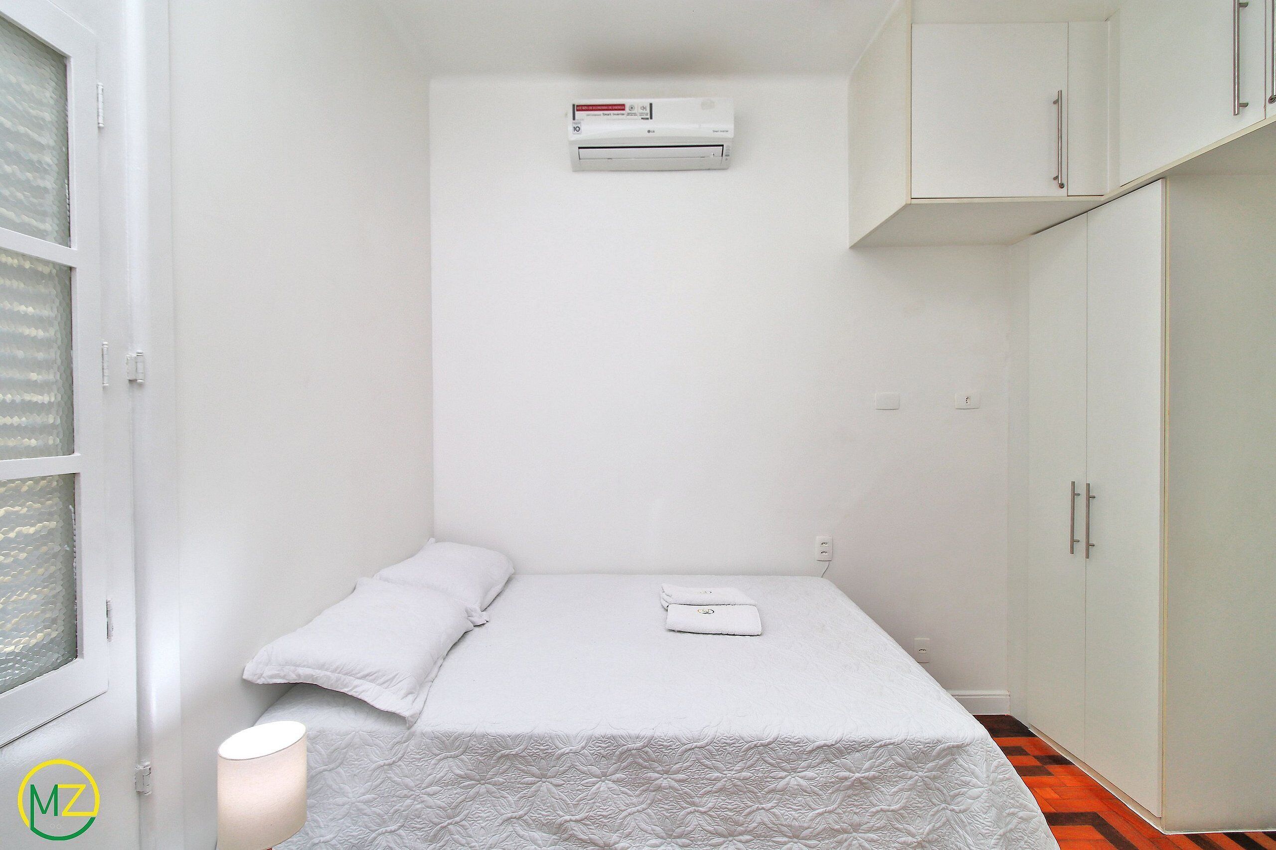 A small, white, single-bed room with a white bedspread, a white pillow, a white bedside table, and a white lamp. There is a white air conditioning unit mounted on the wall above the bed.