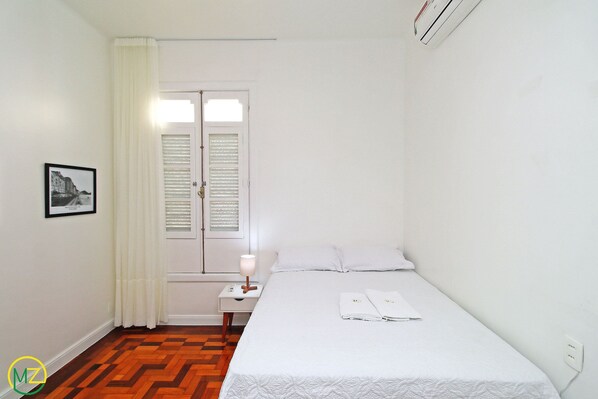A hotel room with a bed, a nightstand, a lamp, a window with blinds, and a framed picture on the wall. - Ronald II (Rio de Janeiro)