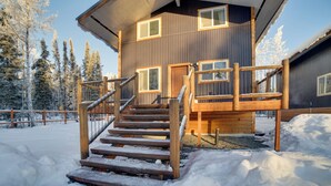 A wooden cabin with a staircase leading to a deck, surrounded by snow-covered trees. - Walk to Kenai River: Forested Fisherman's Cabin! (Sterling)