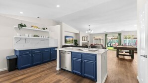 A modern kitchen with blue cabinets, a white island, and a dishwasher. There is a trash can to the left and a dining area with a table and chairs in the background. - Twilight Tunes - Pool, Glowing Arcade Experience (Indio)