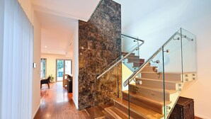 A modern staircase with glass railings and wooden steps, leading to a room with a large window and a plant. - Cozy 3-bedroom house with Wifi/AC and only mins from cafes and perth cbd. (Subiaco)