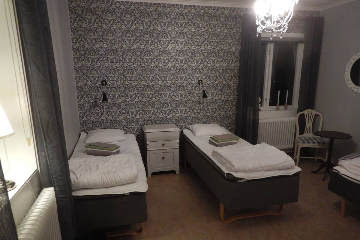Quarto com duas camas de solteiro, uma mesinha, uma cadeira, radiador e papel de parede estampado.