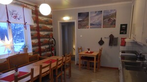 A cozy dining area with wooden tables and chairs, a kitchen with a sink and stove, and a window with a view of trees. - Jokkmokks Vandrarhem Åsgård (Jokkmokk)