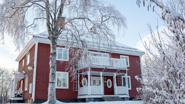 A two-story red house with a white porch and a snow-covered landscape. - Jokkmokks Vandrarhem Åsgård (Jokkmokk)