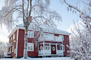 Una casa roja de dos pisos con un porche blanco y un paisaje cubierto de nieve.