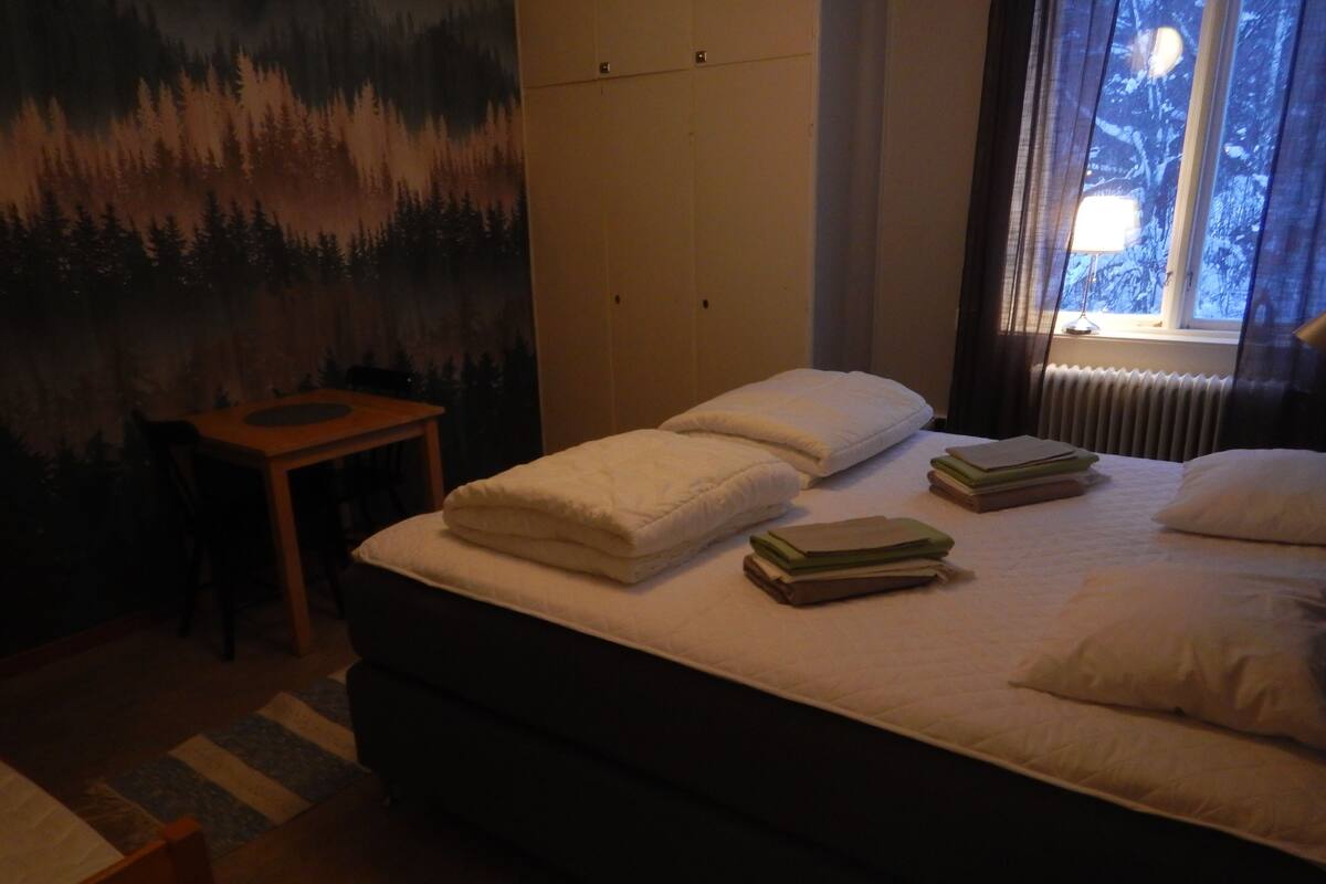 Quarto com cama, almofadas, mesa de cabeceira e janela com vista para árvores.