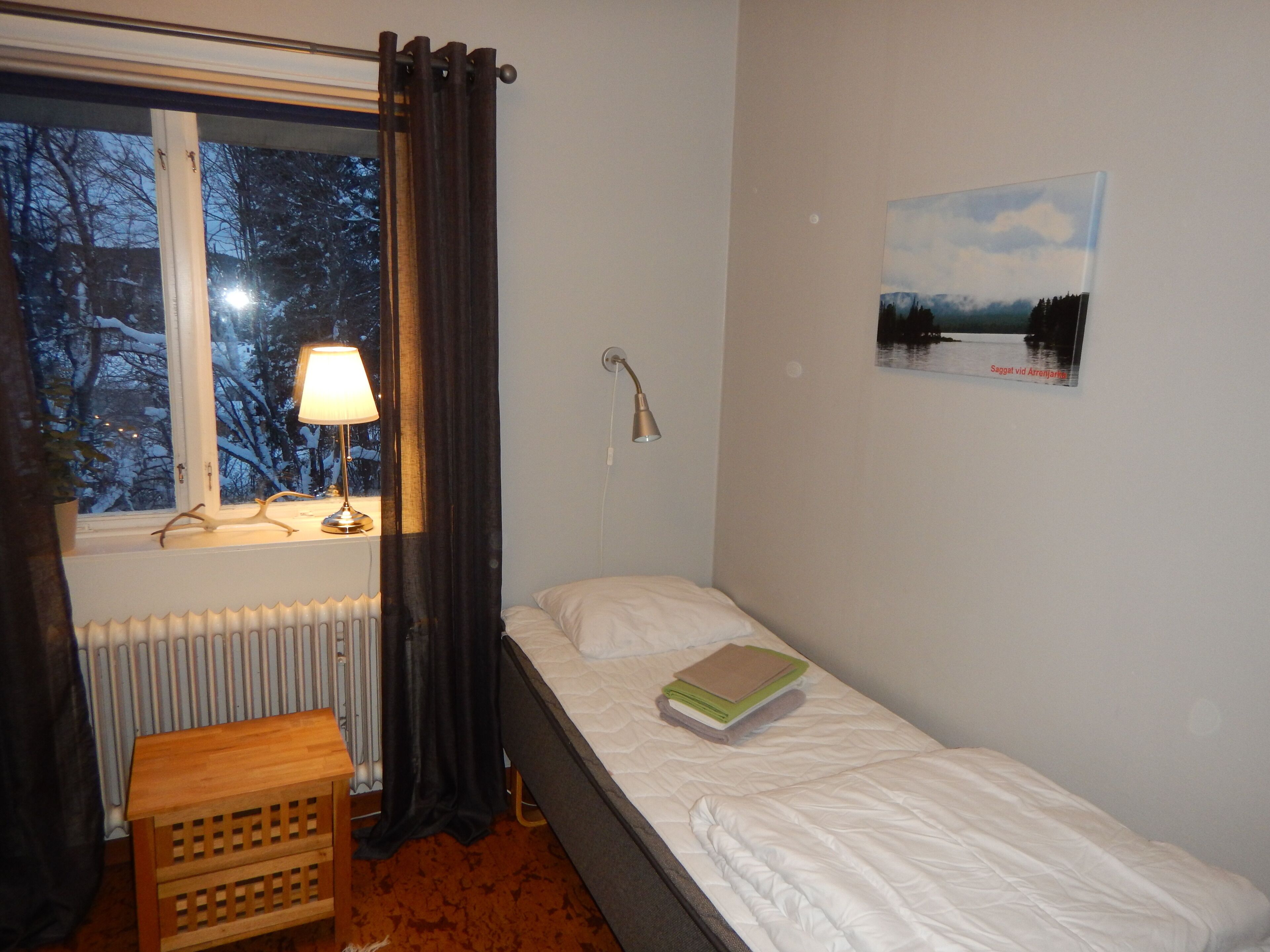 Une petite chambre avec un lit simple, une fenêtre, un radiateur, une table de chevet, une lampe, un tableau au mur et un petit tabouret en bois.