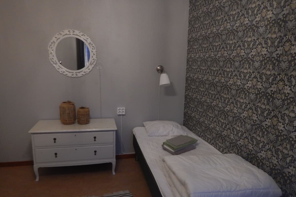 Quarto com cômoda branca, cama com edredom branco, espelho redondo e luminária.