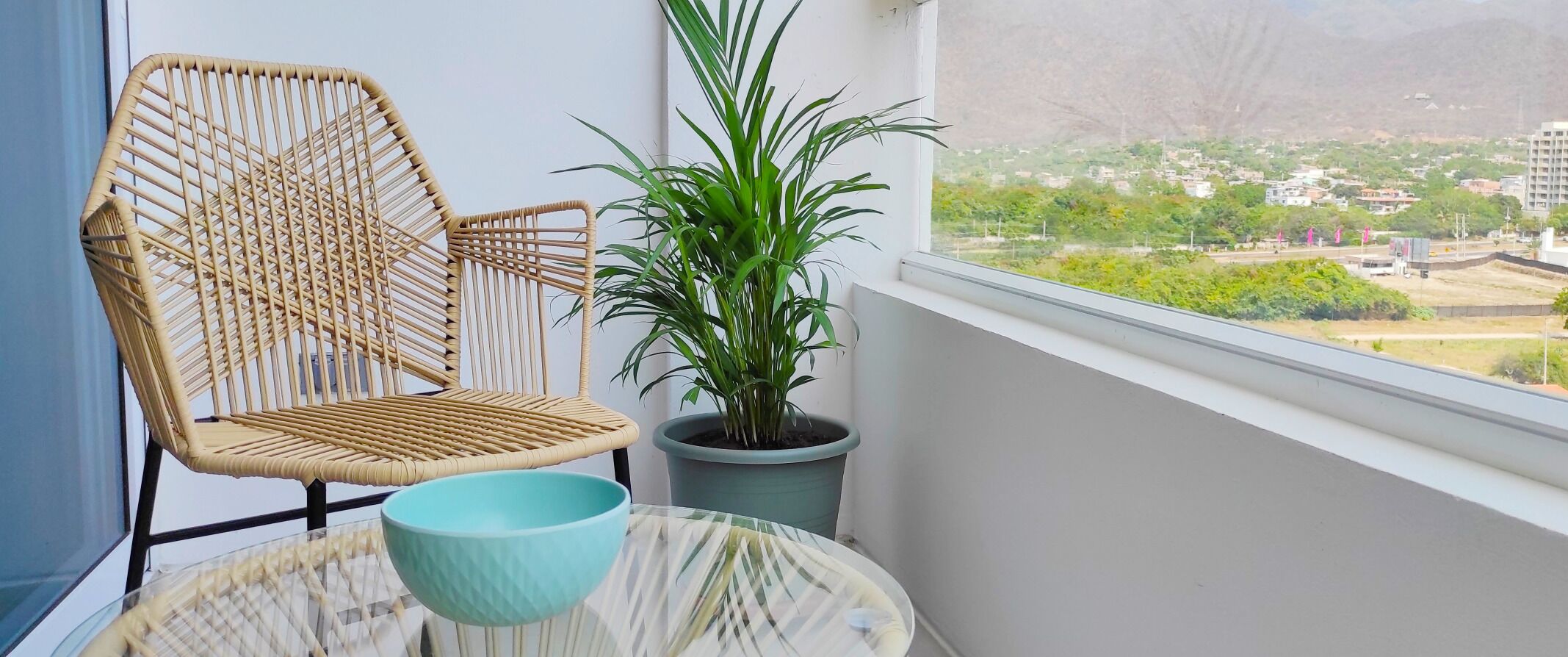 Une chaise en osier, une plante en pot et une table avec un saladier sur un balcon donnant sur la ville.