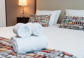 Un lit bien fait, avec des serviettes blanches pliées en rouleau, un coussin à motifs et une lampe sur une table de chevet.