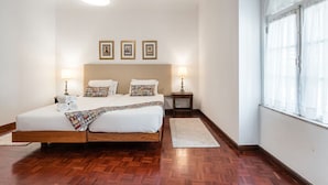 A bedroom with a wooden bed, two bedside tables, a large window with sheer curtains, and three framed pictures on the wall. - Office Residence Six (Oporto)