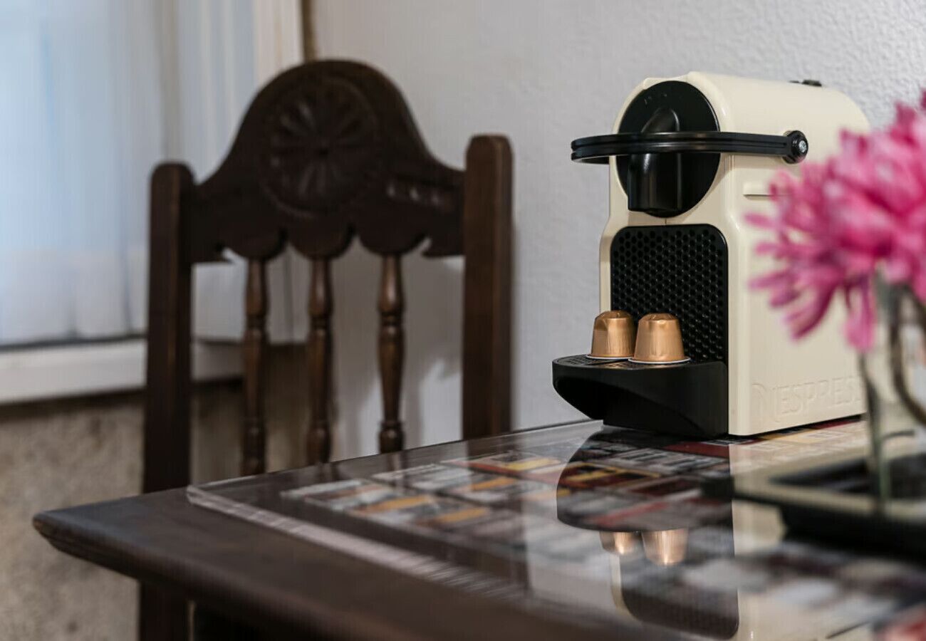 A coffee machine on a table with a magazine, a wooden chair, and a vase with flowers.