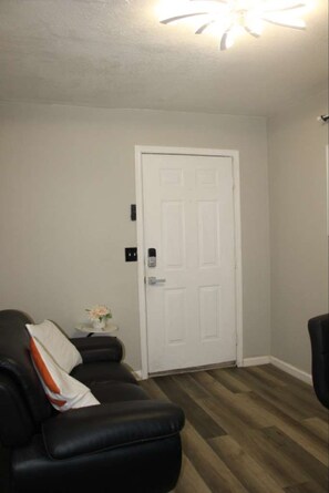 A room with a white door, a black leather sofa with a cushion, a small table with a vase, and a ceiling light fixture.