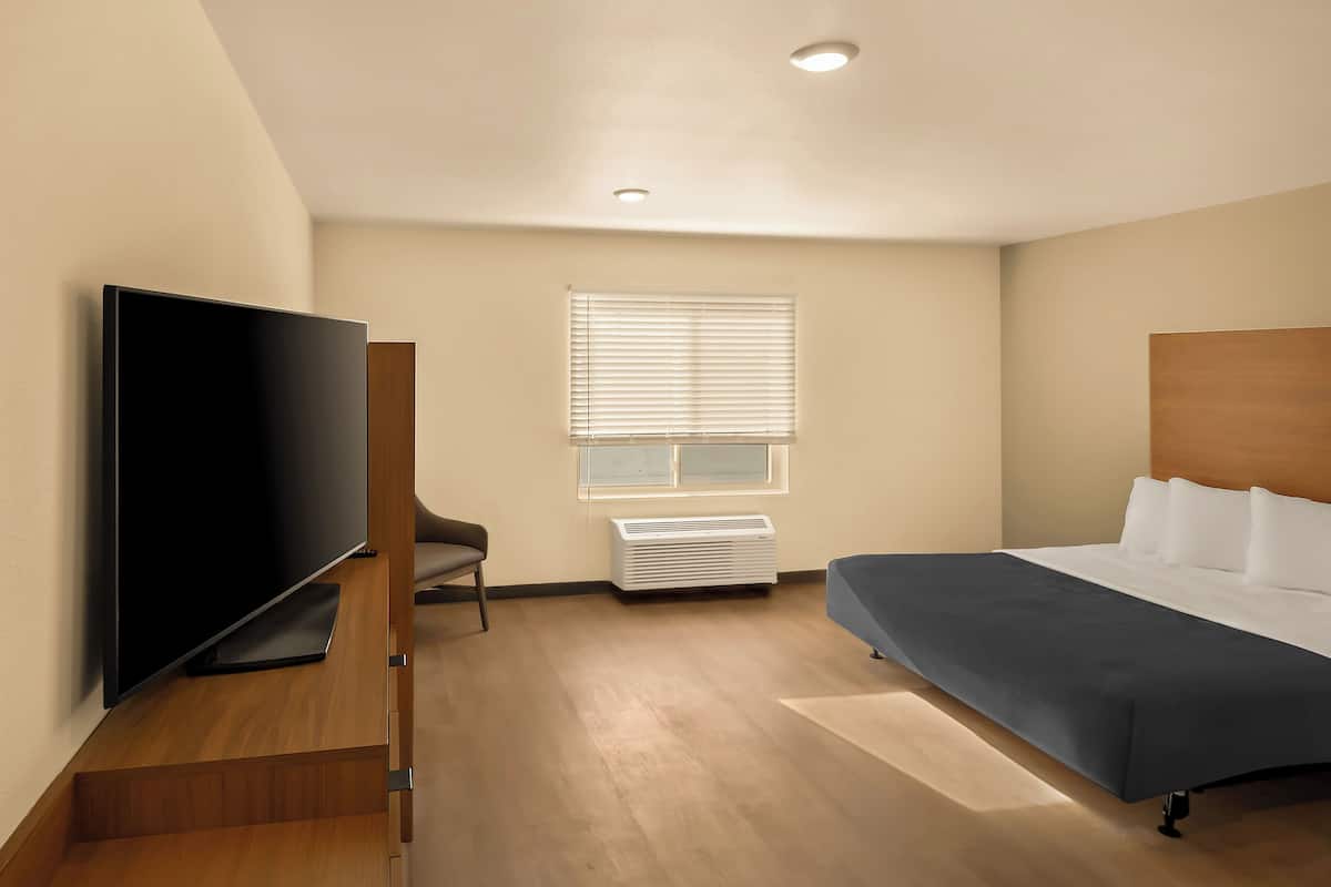 A modern hotel room with a flat-screen TV, a bed, a chair, and a window with blinds.