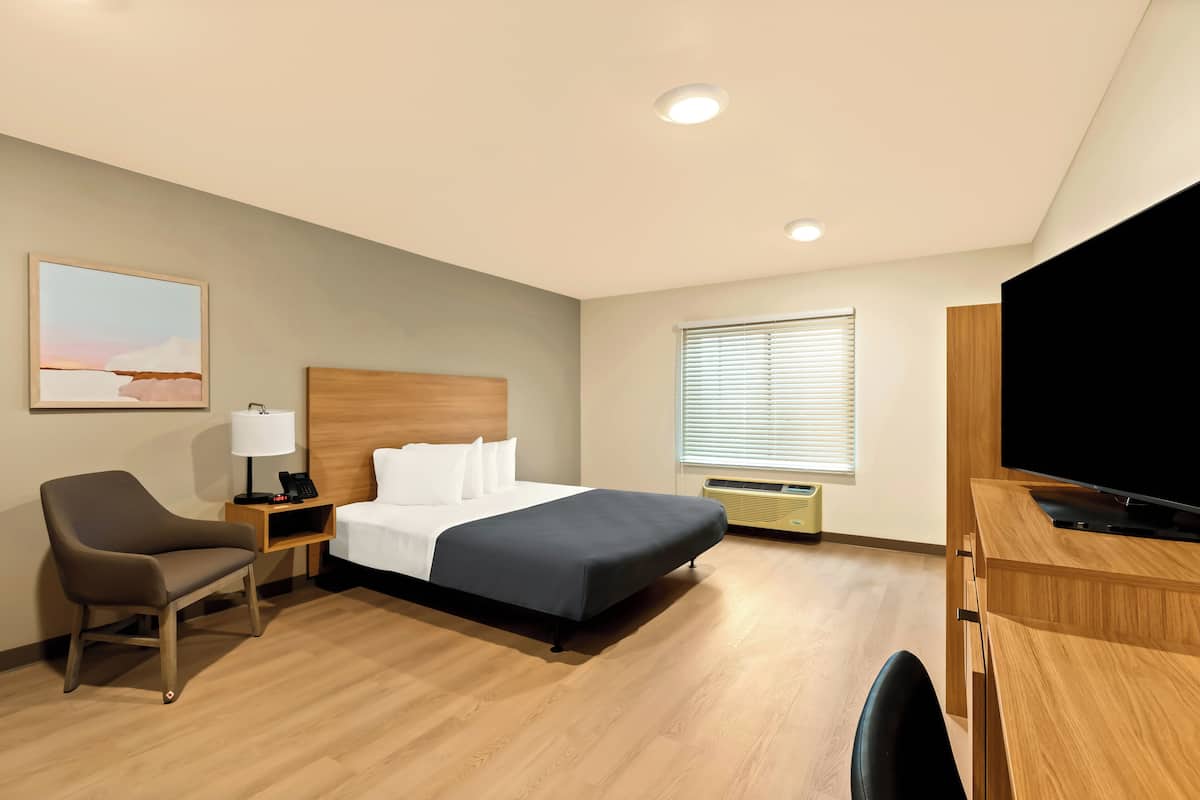 A hotel room with a bed, a desk with a TV, a chair, a bedside table with a lamp, and a window with blinds.