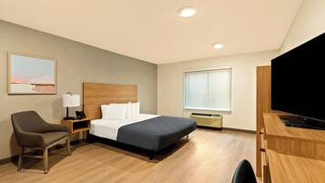 A hotel room with a bed, a desk with a TV, a chair, a bedside table with a lamp, and a window with blinds.