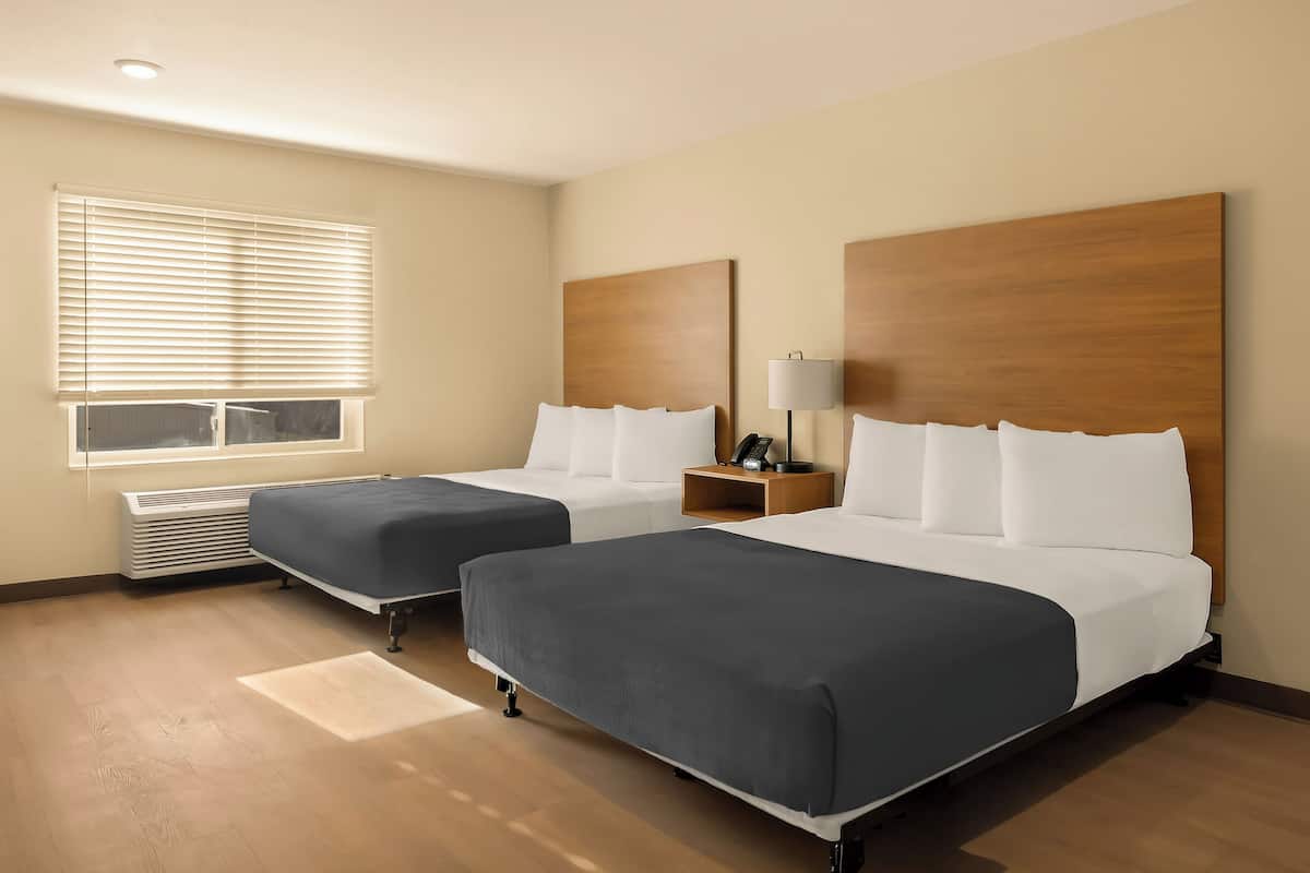 A hotel room with two beds, a window with blinds, a bedside table with a lamp, and a wall-mounted phone.