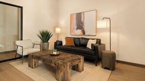 A modern living room with a wooden coffee table, a black leather sofa, a white armchair, a floor lamp, and a large abstract painting on the wall. - LivAway Suites- Syracuse  (Syracuse)