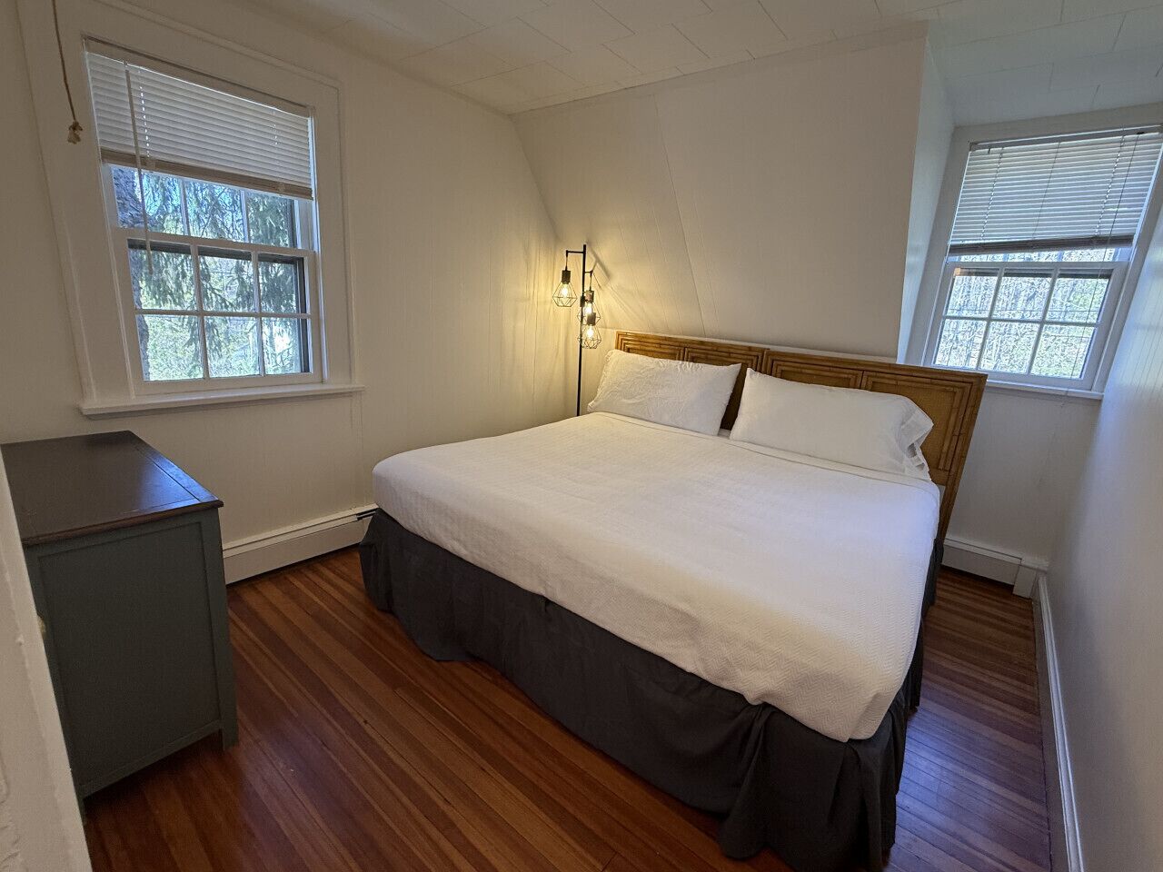 A bedroom with a wooden floor, a bed with white bedding, a nightstand, and a wall-mounted lamp.