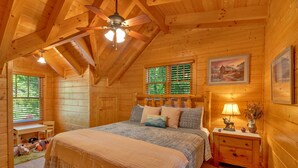 Um quarto de cabana de toras aconchegante, com uma cama grande, um ventilador de teto, uma mesa de cabeceira com abajur e uma janela com persianas.
