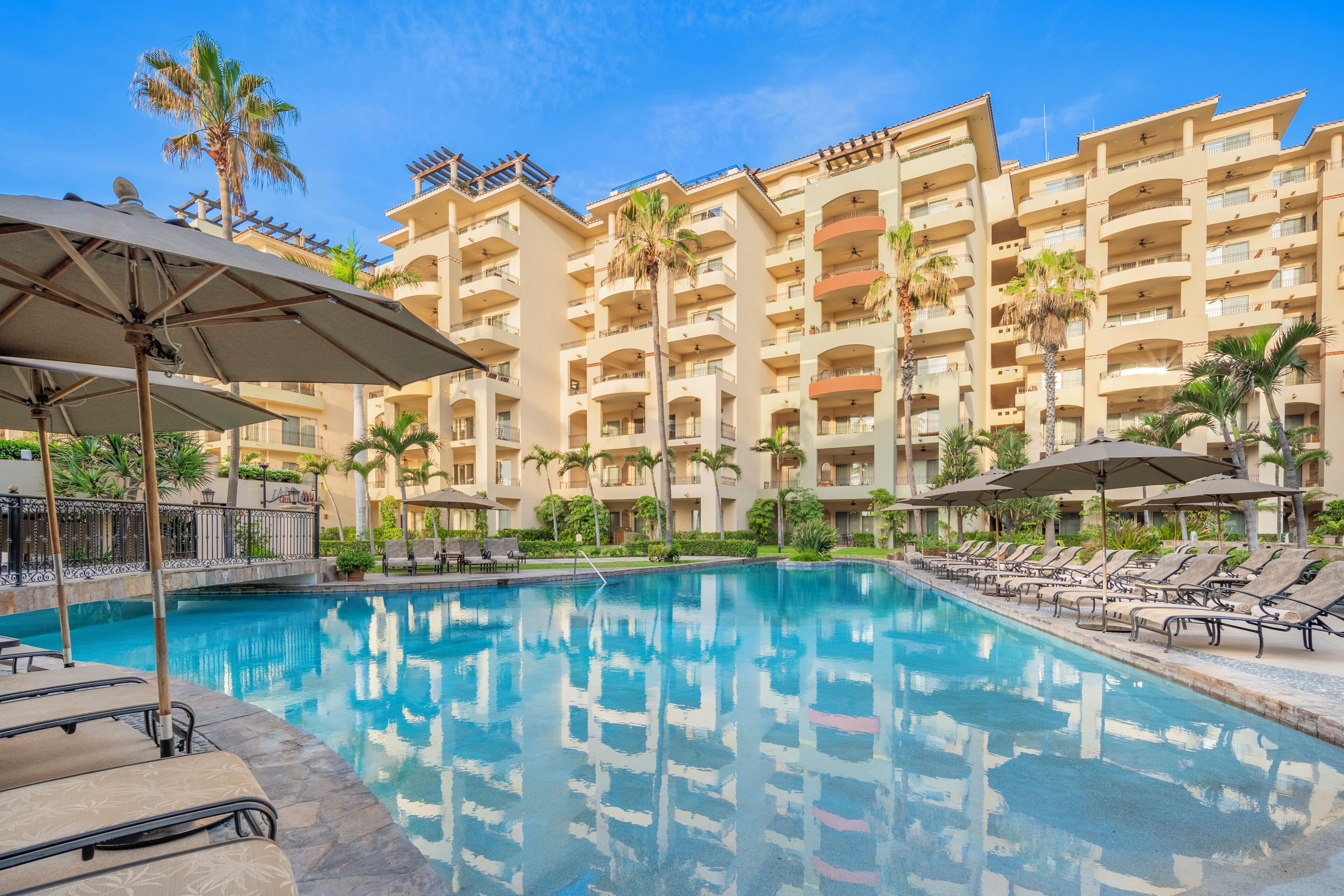 Ein Hotelpoolbereich mit Liegestühlen und Sonnenschirmen, umgeben von Palmen und einem mehrstöckigen Gebäude mit Balkonen.