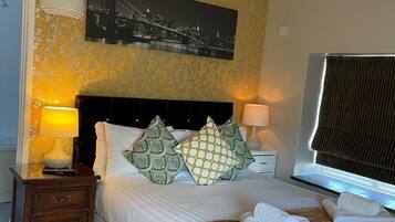 Una cama bien hecha con almohadones estampados, una lámpara de noche y una ventana con persianas.