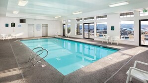 An indoor swimming pool with clear glass walls, a view of the mountains, and a few white chairs around the pool.