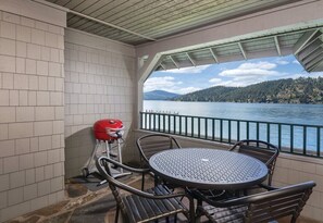 Un balcón techado con mesa y sillas, una parrilla y vistas a un lago y montañas.