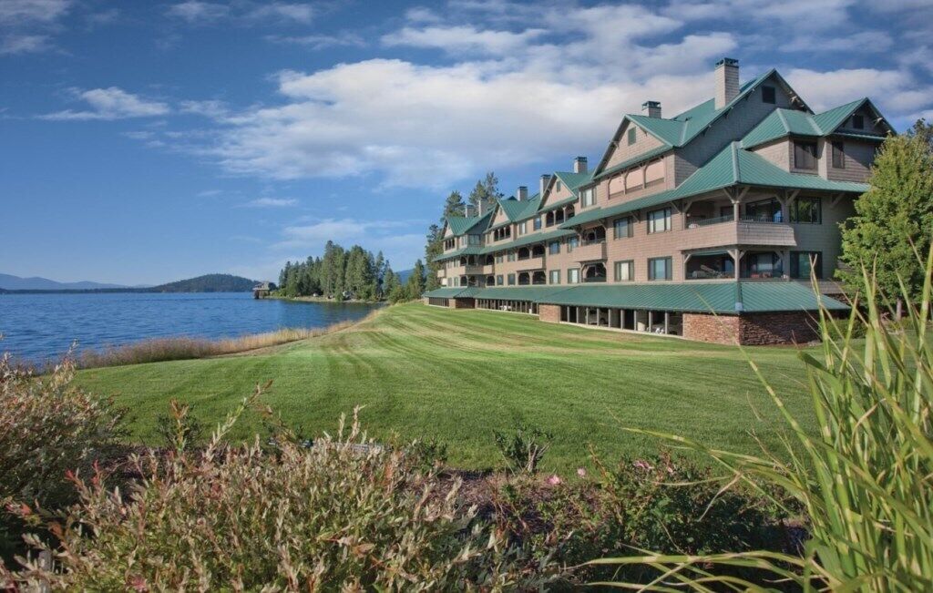 A large, multi-story lodge with a green roof, situated by a lake with a well-maintained lawn in the foreground.