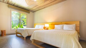 A room with two beds, a window with a view of greenery, and a wooden ceiling.
