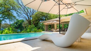 A modern poolside area with a white lounge chair, a large umbrella, and a pool surrounded by lush greenery.