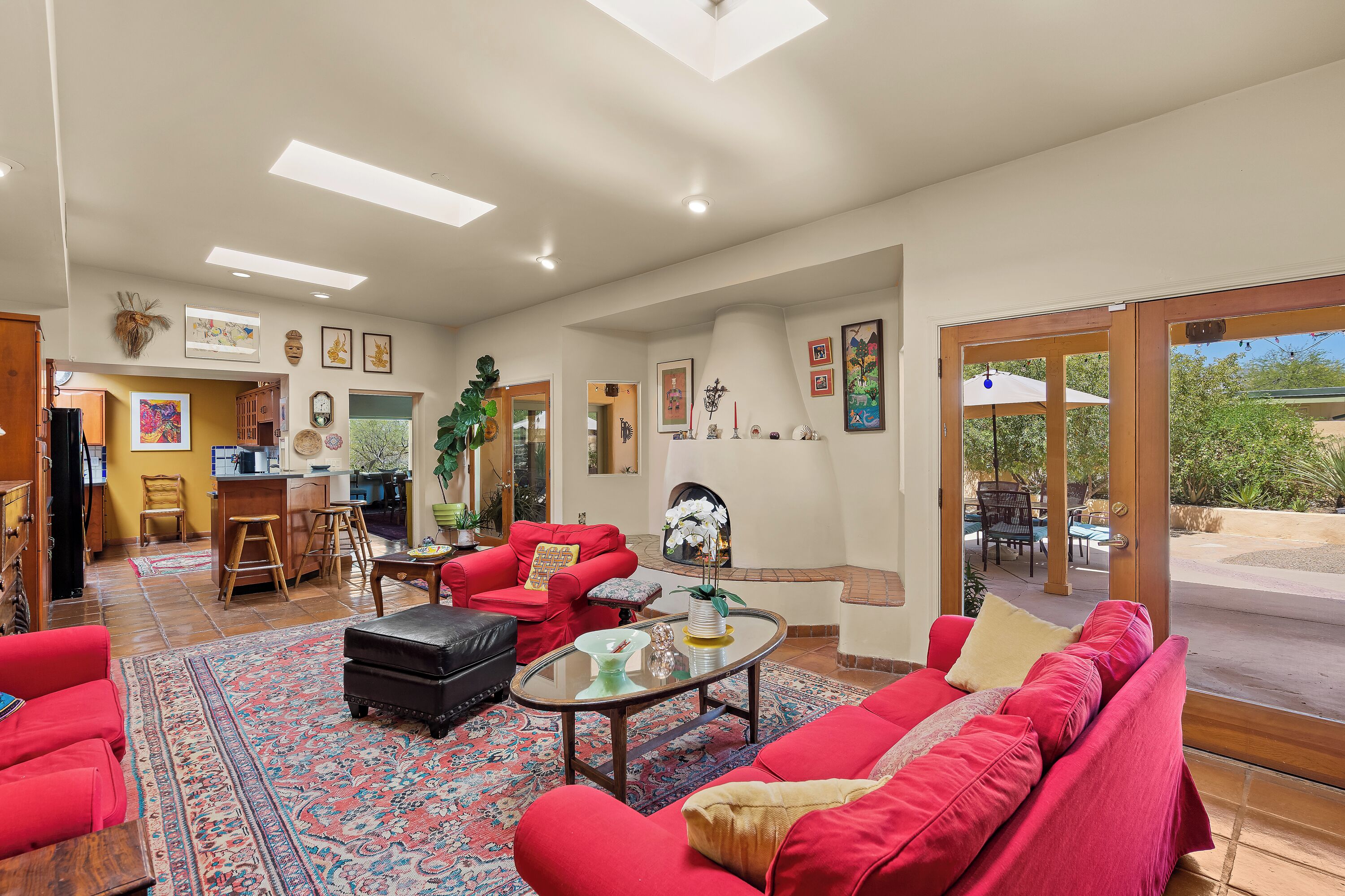 A spacious living room with red sofas, a coffee table, and a large rug. There is a kitchen area in the background with wooden stools and a bar.