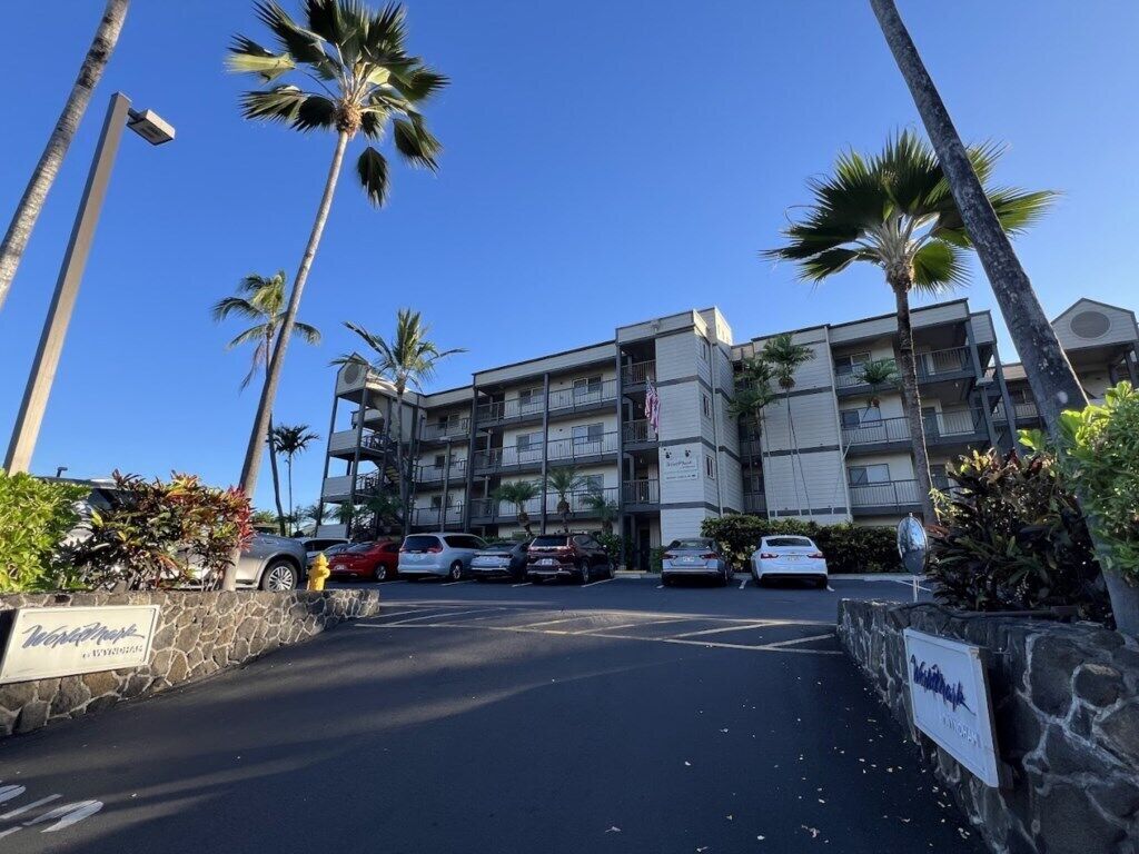 A multi-story hotel with a parking lot, palm trees, and a stone wall with a sign.