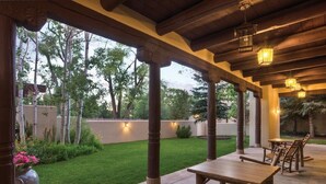 Un patio al aire libre con mesas y sillas de madera, un césped y vistas a árboles y un muro.