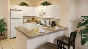 Cocina compacta con armarios blancos, microondas, estufa y un pequeño comedor con una silla y mesa.